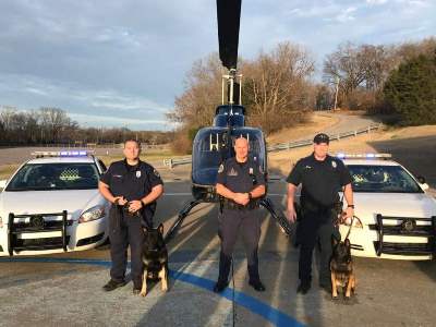Three officers and two K9s standing in front of two police cars and a police helicopter